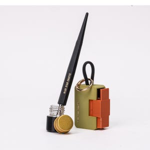 以下が含まれることがあります： 「BLUE STAR CRAFTS」と印刷された黒いペン、金色の蓋が付いた小さなガラス瓶、黒いループと金色のリングが付いた緑とオレンジ色のパズル型キーホルダー。白い背景に配置されています。