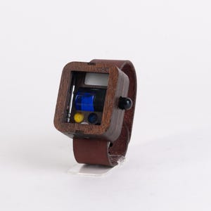 May include: A unique wooden wristwatch with a brown leather band. The watch face is a dark wood square with a black knob and a small display showing blue, yellow, and black components. The watch is displayed on a clear stand.
