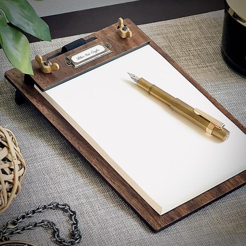 May include: A wooden clipboard with a brass clasp and a blank white notepad. A gold fountain pen is resting on the notepad. The clipboard has a label that reads "Blue Star Crafts".
