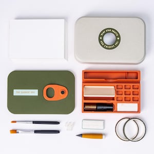 May include: An overhead shot of a craft kit with a variety of art supplies. The kit includes a green box with an orange clasp, an orange tray, paintbrushes, and a stack of white cards. The kit's lid is light gray with a circular design that reads "Blue Star Crafts".