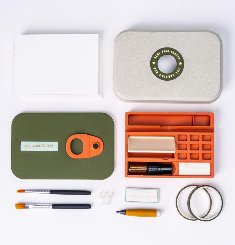 May include: An overhead shot of a craft kit with a variety of art supplies. The kit includes a green box with an orange clasp, an orange tray, paintbrushes, and a stack of white cards. The kit's lid is light gray with a circular design that reads "Blue Star Crafts".