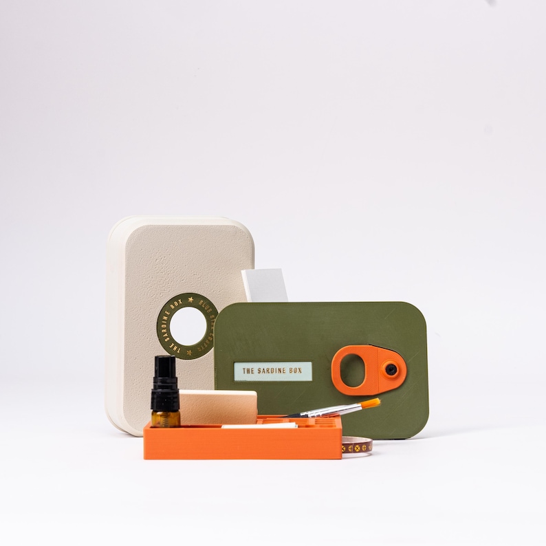 May include: A unique sardine box-shaped kit with a beige container, an olive green box, and an orange tray. The olive green box has an orange handle and the text "THE SARDINE BOX". The kit includes a small bottle and other tools.