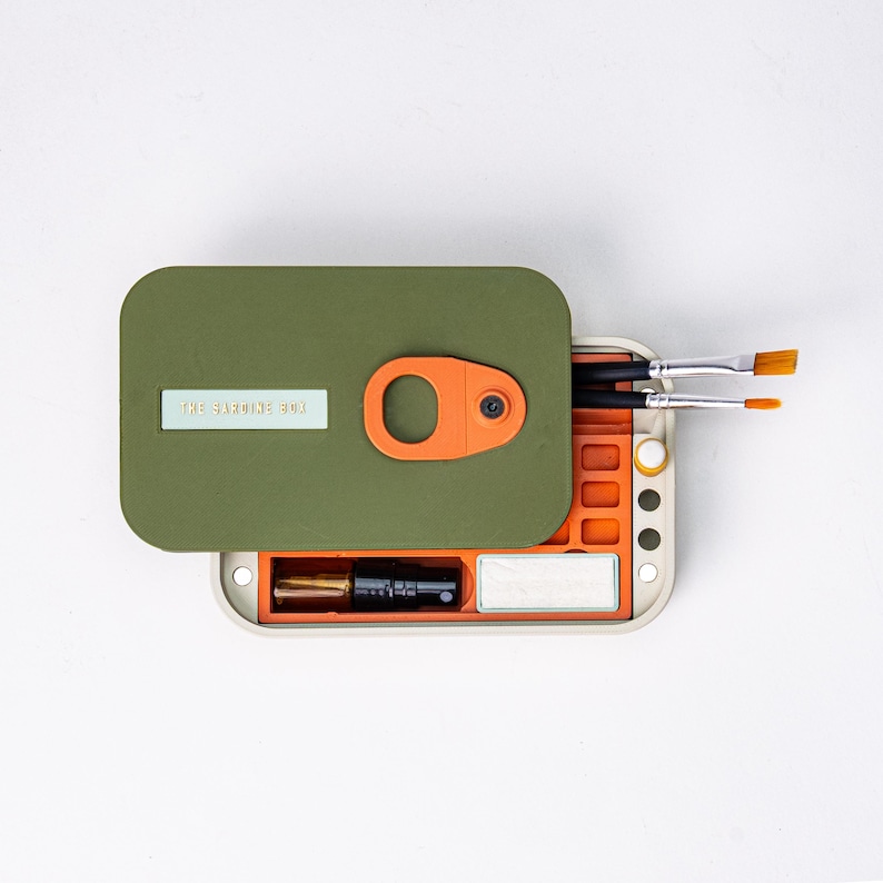 May include: An olive green and orange art supply box with the text "THE SARDINE BOX". The box is open, revealing paintbrushes, a small bottle, and a white rectangular object. The box has an orange handle.