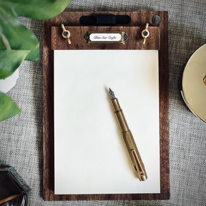 May include: A wooden clipboard with a brass clip and a brass pen holder. The clipboard has a white paper pad and a label that says "Blue Star Crafts". A gold fountain pen is lying on the paper.