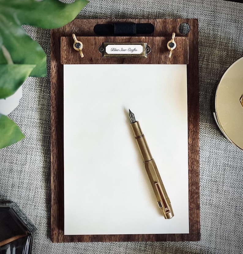 May include: A wooden clipboard with a brass clip and a brass pen holder. The clipboard has a white paper pad and a label that says "Blue Star Crafts". A gold fountain pen is lying on the paper.