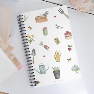Puede incluir: Una libreta de espiral blanca con una ilustración de acuarela de herramientas de jardinería, plantas y flores. La libreta tiene el texto "Melina White Art" en la parte inferior.