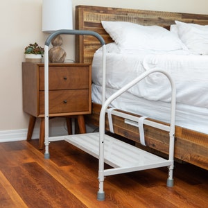 May include: White metal bed assist handle with a step stool. The handle has a gray padded grip and is attached to the side of a bed. The step stool has a white metal frame and a white plastic step.