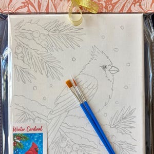 May include: A paint-by-number kit featuring a canvas with a line drawing of a cardinal bird on a pine branch, two blue-handled paintbrushes, and a small reference card labeled Winter Cardinal showing the finished painting.