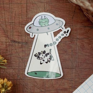 May include: A whimsical sticker featuring a cartoon UFO with a green alien inside, beaming up a black and white cow. The word "mooooo!" is written in a speech bubble. The sticker has a white border and a green base.