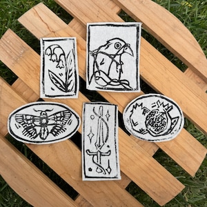 May include: Five fabric patches featuring black ink block-print designs on a light gray background. The designs include a flower, a bird, a moth, a sword with stars, and a pomegranate. The patches are rectangular and oval in shape.