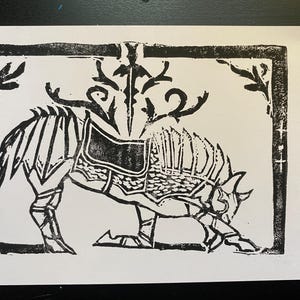 May include: Black and white linocut print of a stylized creature, possibly a wolf or dog, in a kneeling position. The animal is adorned with armor and a saddle, and a sword-like element is above its back. The print is framed with decorative elements.