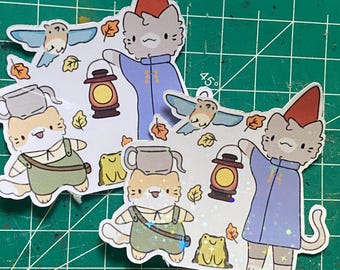May include: Two cat-shaped stickers with a cartoon style. One cat wears a red hat and blue coat, holding a lantern. The other cat wears overalls and a white shirt. Both stickers have autumn-themed elements.