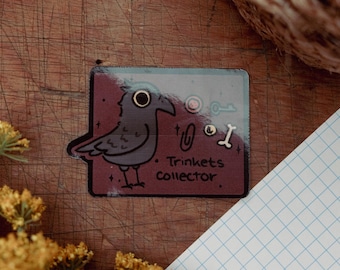 May include: A rectangular sticker featuring a cartoon raven with the text "Trinkets collector." The raven is gray with a black beak and eye. The sticker also includes illustrations of a key, a paperclip, and a bone.