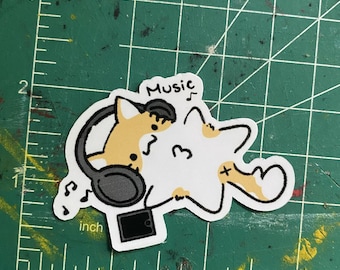 May include: A sticker featuring a cartoon fox wearing headphones and listening to music. The fox is tan and white, with a black outline. The word "Music" is written above the fox's head. Musical notes are scattered around the fox.