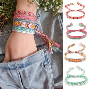 以下が含まれることがあります： カラフルな友情ブレスレットのコレクション。ブレスレットは、ピンク、緑、黄色、白の色合いで複雑な織り模様が特徴です。手首に巻かれたものもあれば、別々に表示されているものもあります。背景にはデニムジャケットと花が含まれています。