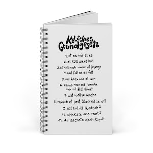 May include: A white notebook with a black spiral binding. The cover features the text "Kölsches Grundgesetz" in a stylized font. The notebook lists eleven rules in German, each starting with a number.