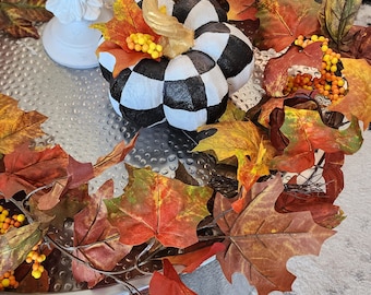Op de afbeelding: Een decoratief herfstarrangement met een zwart-witte geblokte pompoen met een gouden steel, omringd door kleurrijke herfstbladeren en bessen. De opstelling staat op een zilveren dienblad.