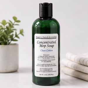 May include: A green bottle of concentrated mop soap with a black cap. The label reads "Clean Cotton" and "Maggie's Farm & Aromatics." The bottle contains 16 fl oz (473.20 g) of soap. A small plant in a white pot is in the background.