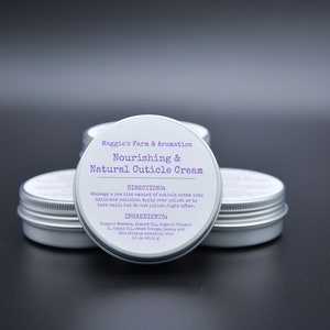 May include: Four small, silver-colored tins of cuticle cream are arranged on a dark surface. Each tin has a white label with purple text that reads "Nourishing & Natural Cuticle Cream" and includes directions and ingredients.