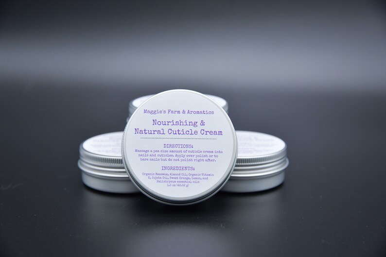 May include: Four small, silver-colored tins of cuticle cream are arranged on a dark surface. Each tin has a white label with purple text that reads "Nourishing & Natural Cuticle Cream" and includes directions and ingredients.