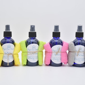 May include: Four blue bottles of yoga mat cleaner with lavender and tea tree essential oils, each wrapped in a different colored microfiber cloth: yellow, green, pink, and gray. The bottles are labeled "Yoga Mat Cleaner" with the brand name "Maggie's Farm & Aromatics".