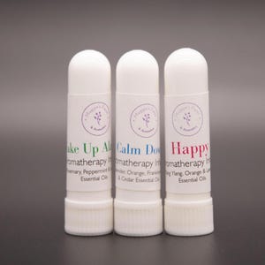 May include: Three white cylindrical aromatherapy inhalers with labels featuring the Maggie's Farm & Aromatics logo. The labels read Wake Up Alert with green text, Calm Down with blue text, and Happy with pink text, listing essential oil blends.