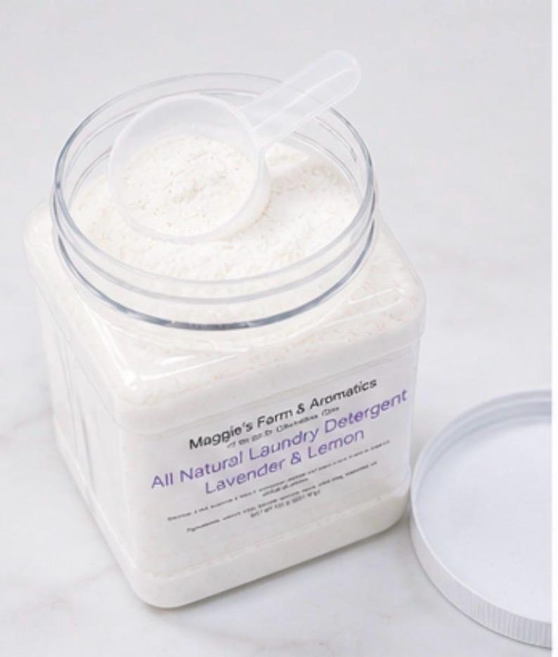 Puede incluir: Un recipiente cuadrado transparente de detergente blanco para ropa con una cuchara a juego encima. La etiqueta dice "All Natural Laundry Detergent Lavender & Lemon". La tapa est&aacute; a un lado.