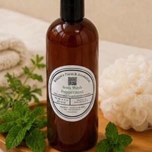 May include: A brown bottle of body wash with a black cap and a white label that reads "Body Wash Peppermint." Fresh mint leaves and a loofah are on a wooden tray. The bottle is surrounded by fresh mint leaves and a white loofah.