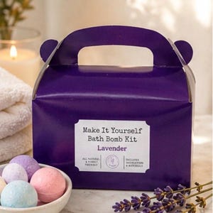 May include: A purple box with a handle and the text "Make It Yourself Bath Bomb Kit Lavender." A small white bowl holds several colorful bath bombs. A sprig of lavender and a lit candle are in the background.