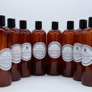 May include: A collection of brown plastic bottles with black caps, each labeled with a white and blue circular sticker. The labels read "Nourishing Body Wash" with various scents like Rose, Peppermint, and Lavender & Vanilla. The bottles are arranged against a white background.