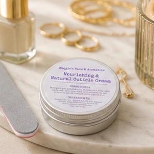 May include: A silver tin of "Nourishing & Natural Cuticle Cream" sits on a marble surface, next to a nail file and a bottle of nail polish. Gold rings, a necklace, and a candle holder are in the background. The cream's label includes directions and ingredients.