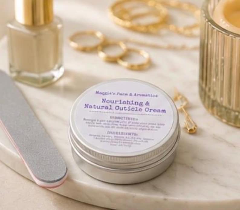 May include: A silver tin of "Nourishing & Natural Cuticle Cream" sits on a marble surface, next to a nail file and a bottle of nail polish. Gold rings, a necklace, and a candle holder are in the background. The cream's label includes directions and ingredients.