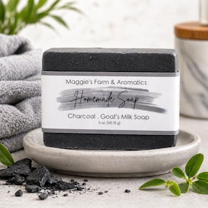 May include: A dark gray, rectangular bar of homemade charcoal and goat's milk soap. The soap is labeled with the text "Maggie's Farm & Aromatics Homemade Soap" and weighs 5 oz. The soap sits on a small, light gray dish, with charcoal pieces and greenery nearby.