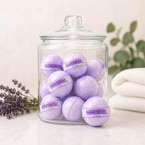 May include: A clear glass jar filled with lavender-scented bath bombs. The bath bombs are light purple and labeled "Handmade Bath Bomb" and "Lavender." A bundle of lavender and a folded white towel are in the background.