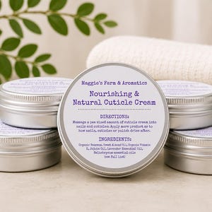 May include: A collection of small, silver metal tins of cuticle cream. The central tin displays the product name, "Nourishing & Natural Cuticle Cream," along with directions and ingredients. The background includes a white towel and green leafy sprigs.