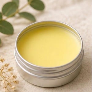 May include: A small, open, silver-colored tin filled with a smooth, pale yellow substance. The tin is round with a ridged exterior. The background includes green leaves and small, dried flowers, suggesting a natural product.