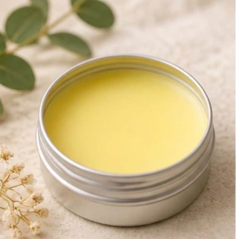 May include: A small, open, silver-colored tin filled with a smooth, pale yellow substance. The tin is round with a ridged exterior. The background includes green leaves and small, dried flowers, suggesting a natural product.