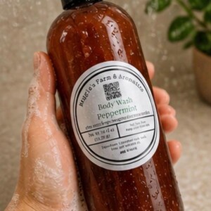 May include: A brown body wash bottle with a black cap is held in a soapy hand. The label reads "Peppermint Body Wash" from "Haugle's Farm & Aromatics". The bottle is wet with water droplets, suggesting use in a shower.