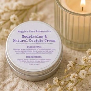 May include: A silver tin of "Nourishing & Natural Cuticle Cream" with text, next to a lit candle in a glass holder and sprigs of white flowers. The cream's label includes directions and ingredients.