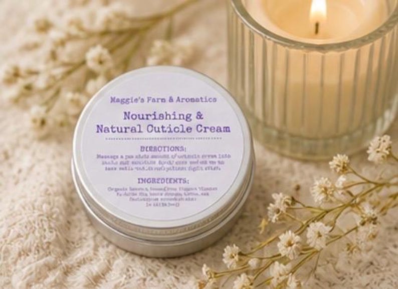 May include: A silver tin of "Nourishing & Natural Cuticle Cream" with text, next to a lit candle in a glass holder and sprigs of white flowers. The cream's label includes directions and ingredients.