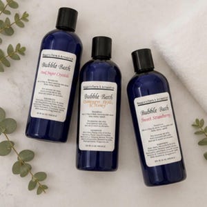 May include: Three dark blue bubble bath bottles with black caps and white labels. The labels read "Bubble Bath" with different scents: "Opal Sugar Crystals", "Orange & Honey", and "Sweet Strawberry". Eucalyptus sprigs and a white towel are in the background.