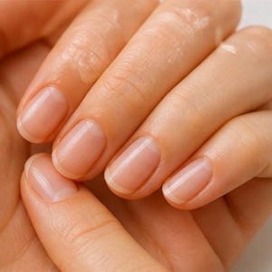May include: Close-up of a hand with well-manicured nails. The nails are a natural, pale pink color with a glossy finish. The skin is smooth and appears moisturized, with a slight sheen. The image focuses on the nails and the texture of the skin.
