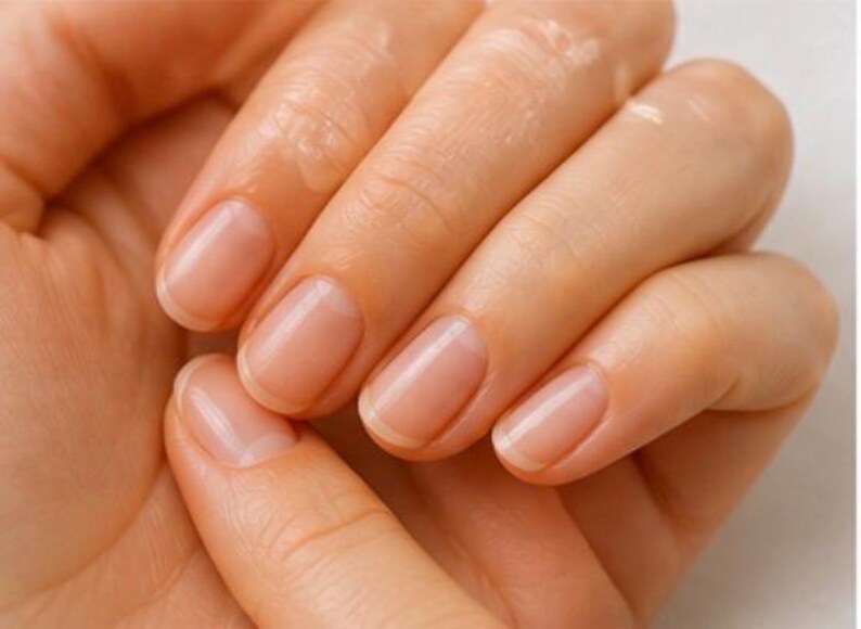 May include: Close-up of a hand with well-manicured nails. The nails are a natural, pale pink color with a glossy finish. The skin is smooth and appears moisturized, with a slight sheen. The image focuses on the nails and the texture of the skin.
