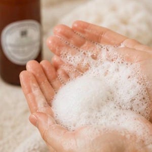 May include: Hands cupping a large amount of white, foamy soap bubbles. A brown bottle with a black cap and a white label is in the background, out of focus. The image suggests a skincare or bath product.