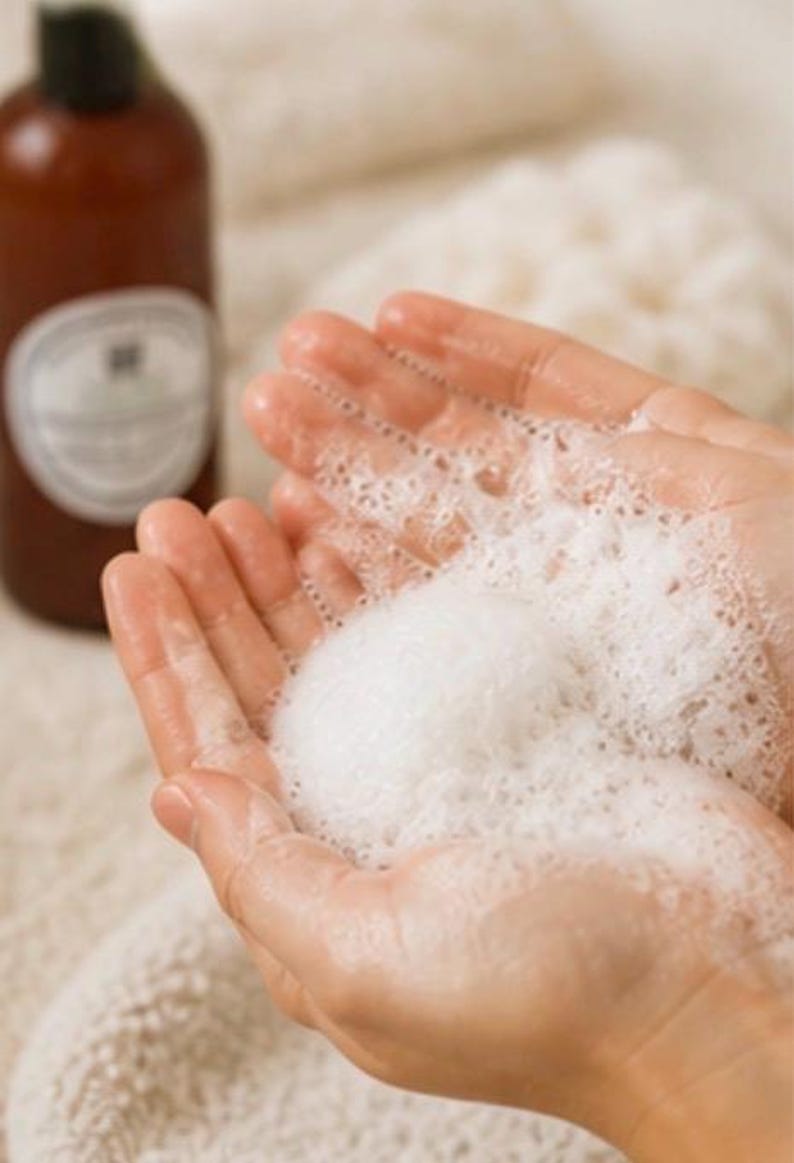 May include: Hands cupping a large amount of white, foamy soap bubbles. A brown bottle with a black cap and a white label is in the background, out of focus. The image suggests a skincare or bath product.