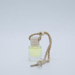 May include: A small, clear glass bottle filled with a pale yellow liquid. The bottle has a light beige cap and is suspended by a beige and gold rope. The product is likely an air freshener or essential oil diffuser.
