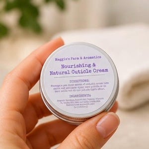 May include: A small, round, silver tin of cuticle cream held in a hand. The label reads "Maggie's Farm & Aromatics, Nourishing & Natural Cuticle Cream." The label also includes directions and ingredients. The background is blurred.