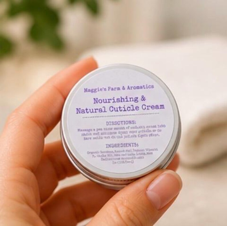 May include: A small, round, silver tin of cuticle cream held in a hand. The label reads "Maggie's Farm & Aromatics, Nourishing & Natural Cuticle Cream." The label also includes directions and ingredients. The background is blurred.