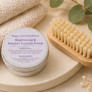 May include: A silver tin of "Nourishing & Natural Cuticle Cream" sits on a white tray next to a wooden nail brush and folded white towels. The cream's label includes the brand name "Maggie's Face & Aromatics". The brush has light-colored bristles.
