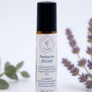 May include: A roll-on bottle labeled "Headache Relief" with a black cap and a white label. The label includes the brand name, product name, and ingredients. The bottle is surrounded by lavender and greenery.